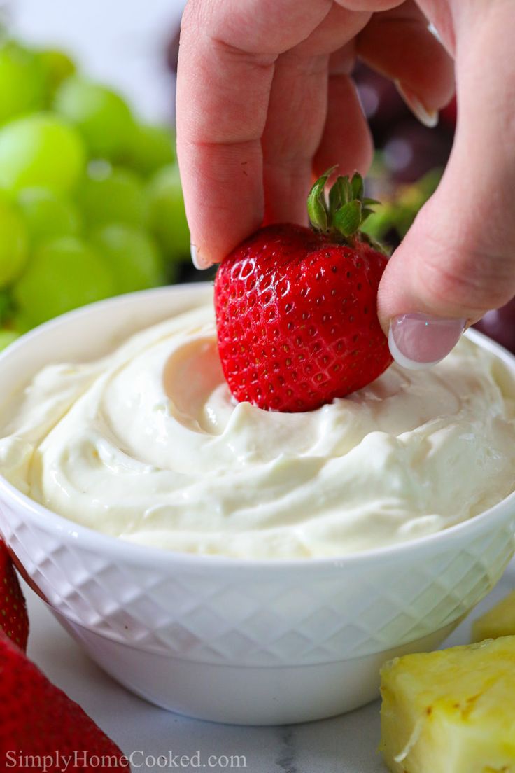 a hand dipping a strawberry into a bowl of cream cheese with grapes in the background