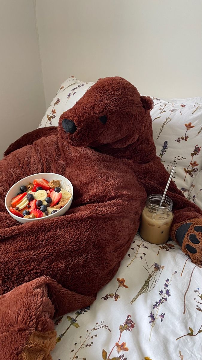 a brown teddy bear laying on top of a bed next to a bowl of fruit