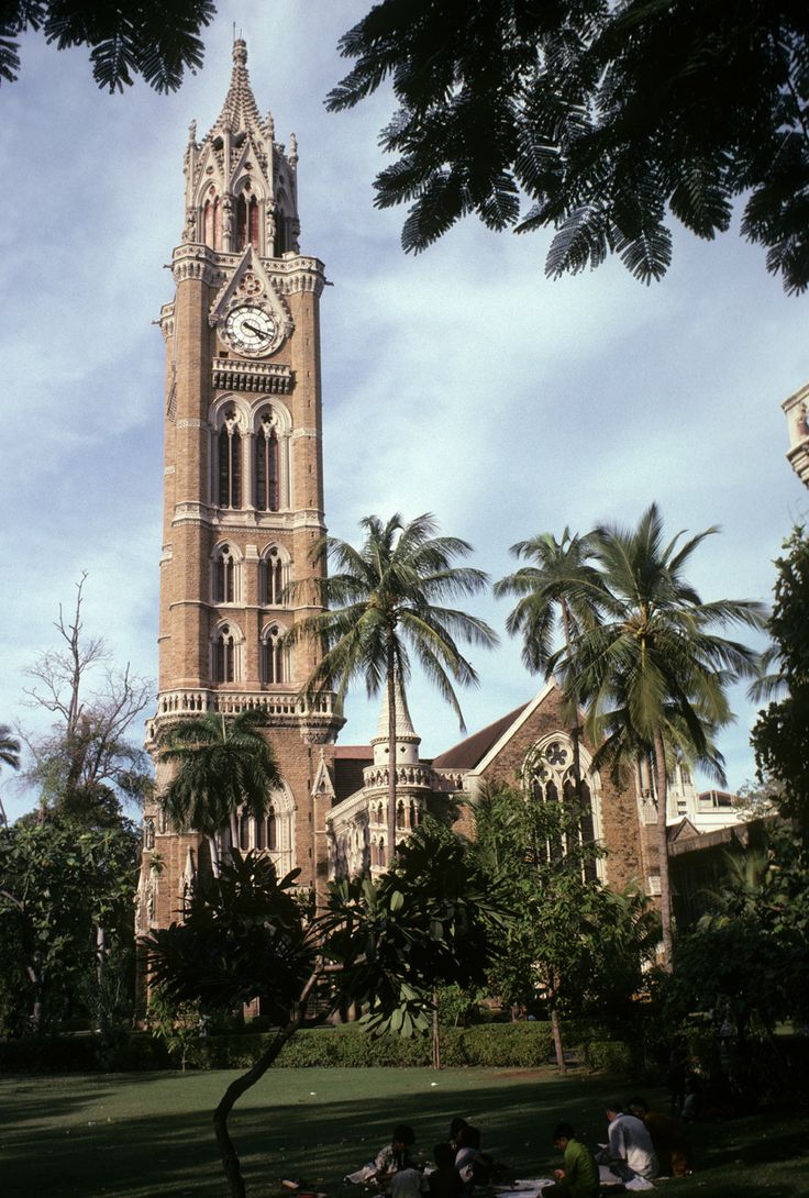 people are sitting on the grass in front of a tall building with a clock tower