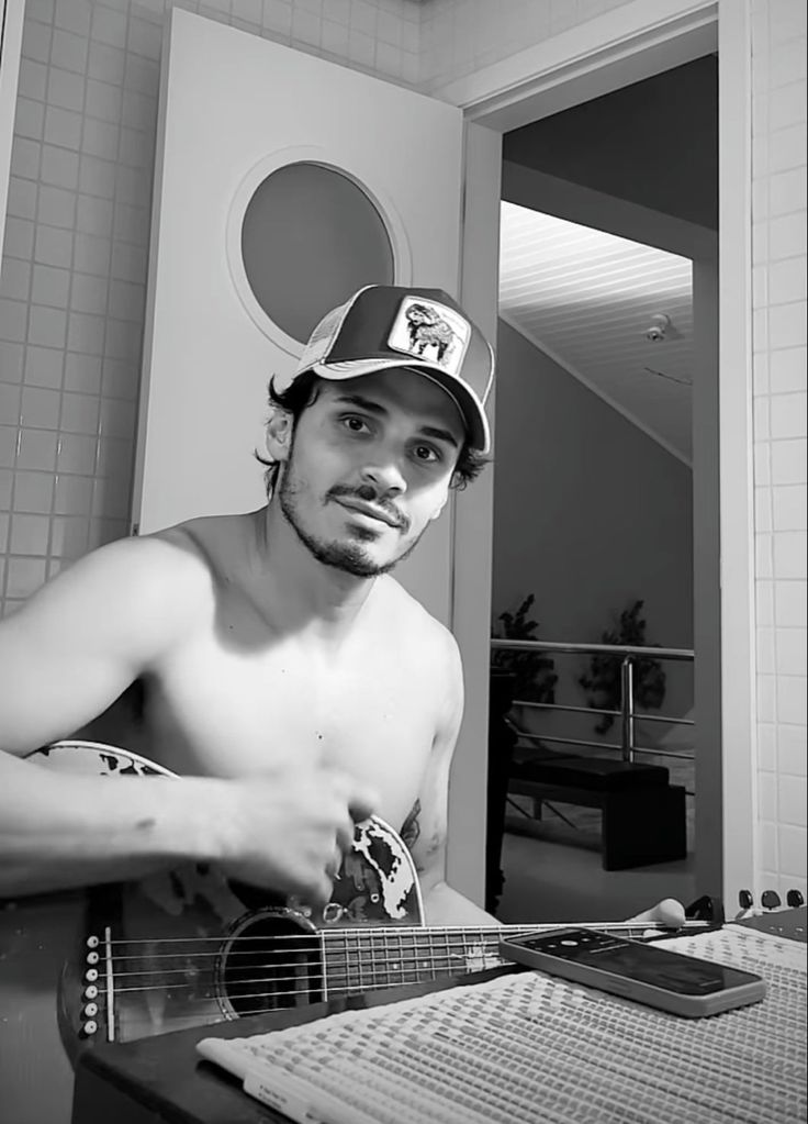 a shirtless man holding an ukulele and looking at the camera while wearing a baseball cap