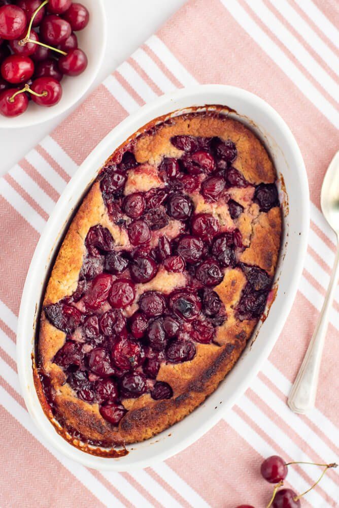 a cake with cherries in it on a pink and white table cloth next to bowls of cherries