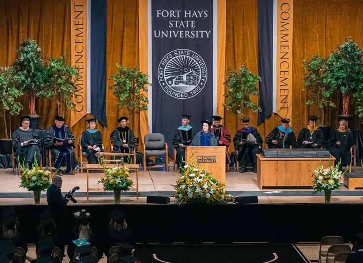 people are sitting on stage in front of an audience at a graduation ceremony with trees and flowers