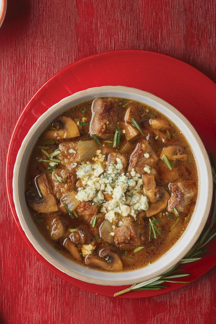 a bowl of soup with meat, mushrooms and cheese on a red plate next to a spoon