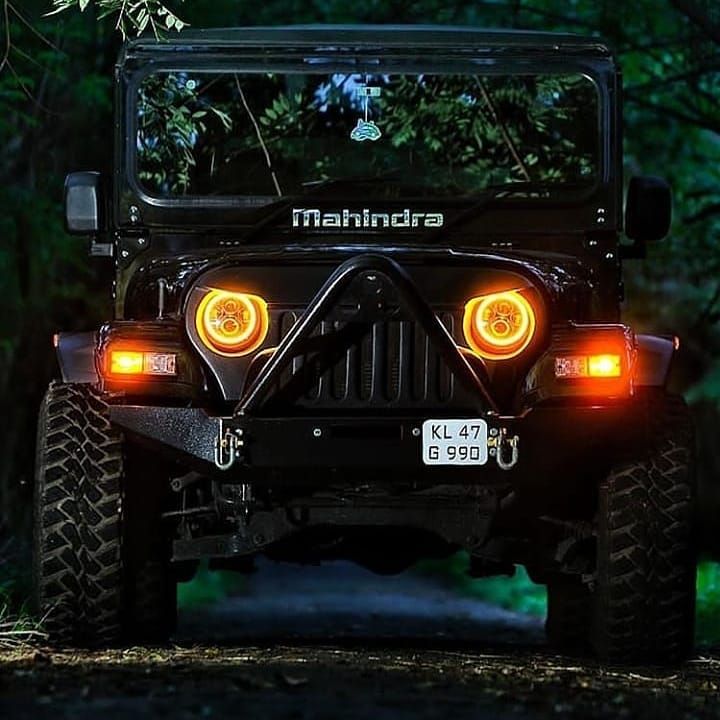 the front end of a black jeep with its lights turned on and trees in the background