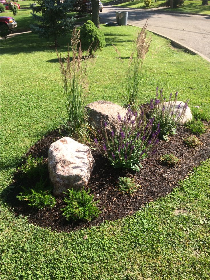 a rock sitting in the middle of a flower bed on top of grass next to a road