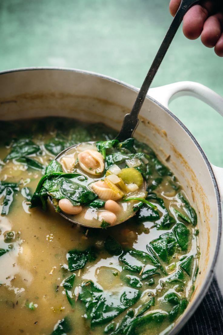 a person holding a ladle full of soup with spinach and other food items