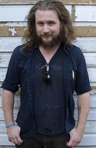 a man with long hair and beard standing in front of a white wall wearing sunglasses