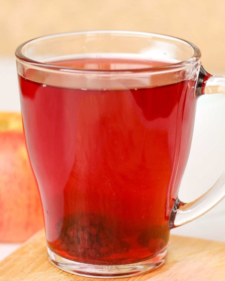 a glass mug filled with red liquid sitting on top of a cutting board next to an apple