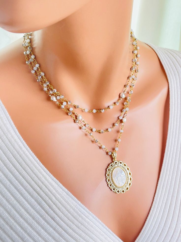 "This is a stunning multi strand necklace made with opaque grey ombre faceted 4mm crystals with scattered pearls, mother of pearl shell etched with Miraculous Mary image surrounded with crystals pendant. This necklace has three layers all connected in the back with a clasp.  The 18k gold filled pendant measures 38 mm x 30 mm and hangs on a 9mm bail. This necklace is absolutely stunning. Comes in a gift box ready to present. Model is wearing 15-to-18-inch length. Top strand is 15\" around the nec White Double Strand Layered Jewelry, White Layered Double Strand Jewelry, White Spiritual Necklaces For Layering, White Spiritual Necklace For Layering, Spiritual White Necklaces For Layering, Elegant Layered Necklace With Faceted Beads For Gift, Elegant Jewelry With Miraculous Medal And Round Beads, Elegant White Miraculous Medal Jewelry, White Necklace With Miraculous Medal And Round Beads