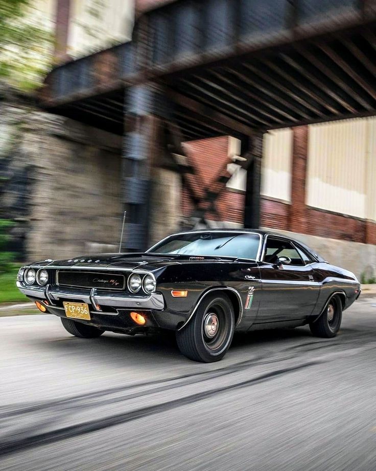 an old black muscle car driving down the street in front of a brick building and bridge