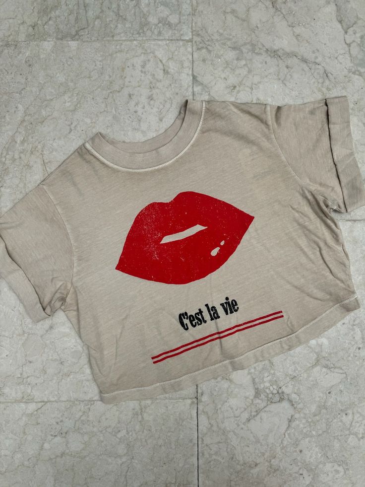 Show off your care free attitude with this Girl Dangerous C'est La Vie Tee with a bold red lips graphic. Perfect for adding a touch of sass to any outfit, this shirt is a must-have for anyone who loves to have fun and live life to the fullest. Color: Sand 100% Cotton Machine wash cold inside out with like colors, tumble dry low Fitted Tan T-shirt With Graphic Print, Cheap Tan T-shirt With Text Print, Pink V-neck Graphic Tee Top, Girl Gang Shirt, Trendy Leopard Print Graphic T-shirt, Bold Red Lips, Swim Shoes, Short Rompers, Board Shorts