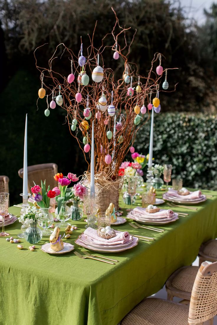 the table is set for easter dinner with green linens and colorful eggs hanging from branches