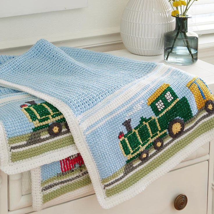 a crocheted train blanket on top of a dresser next to a vase with flowers