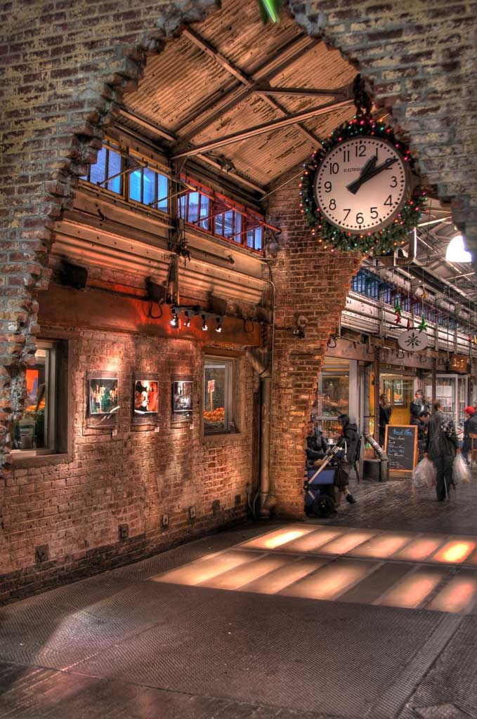 a clock hanging from the side of a brick wall in a train station with people walking by