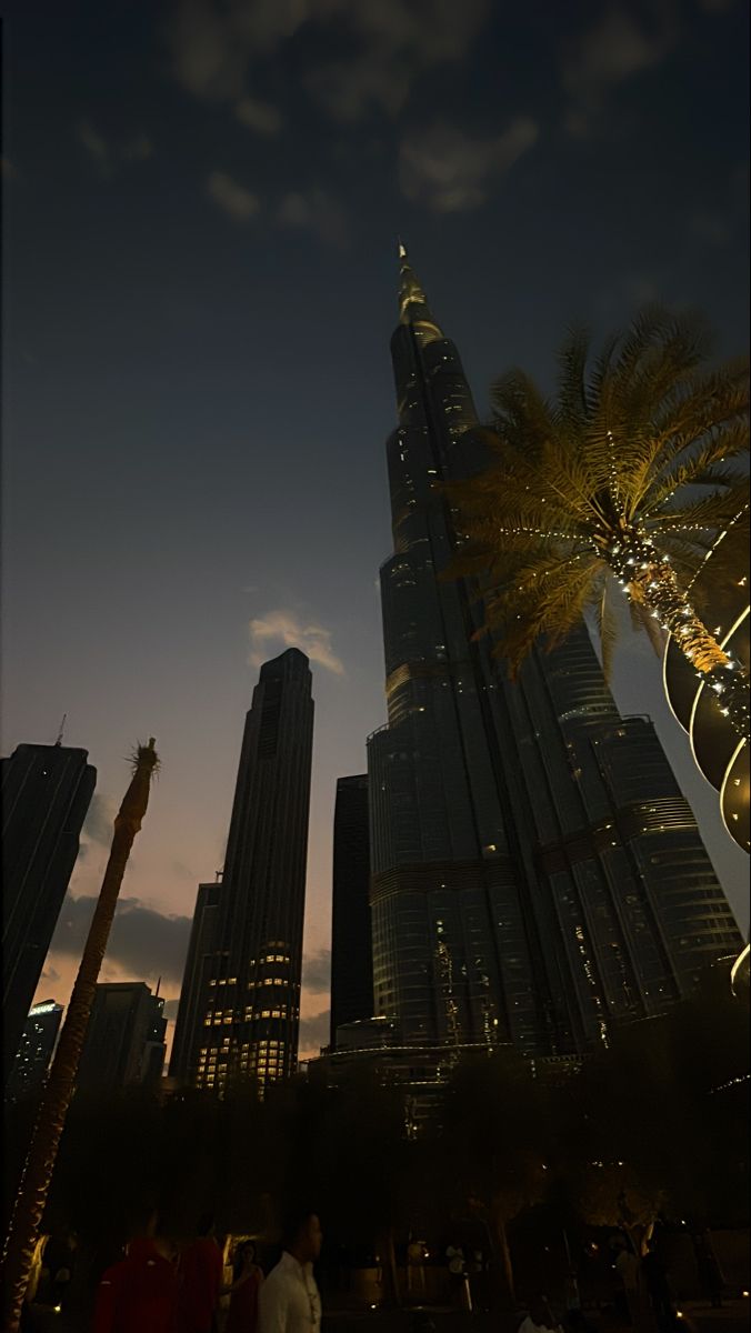 people walking in front of skyscrapers at night with palm trees and lights on them