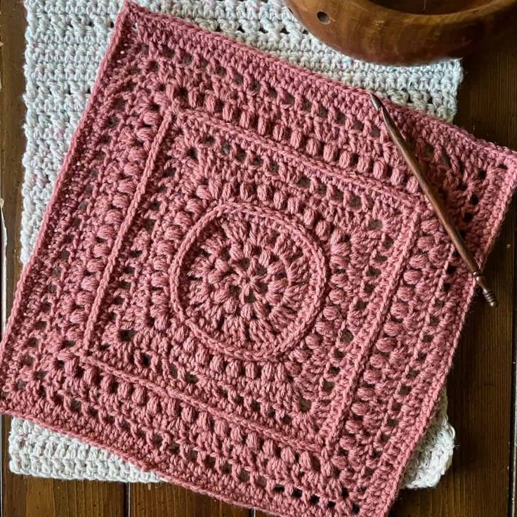 two crocheted squares sitting on top of a wooden table next to a bowl