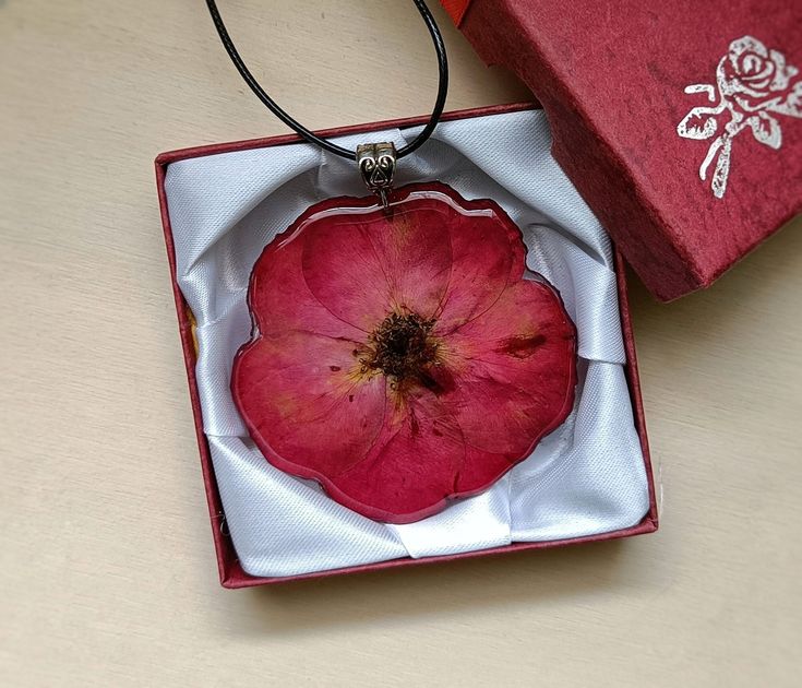 an open red box with a flower inside on a table next to a necklace that is in the shape of a heart