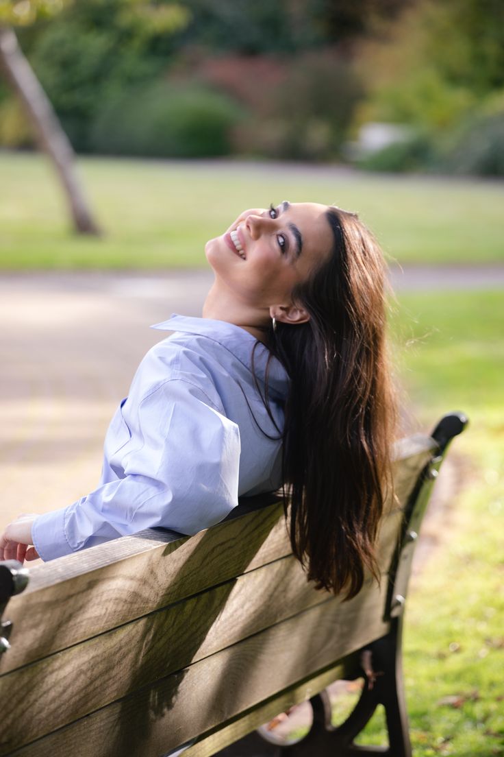 a woman sitting on top of a wooden bench next to a lush green park filled with trees