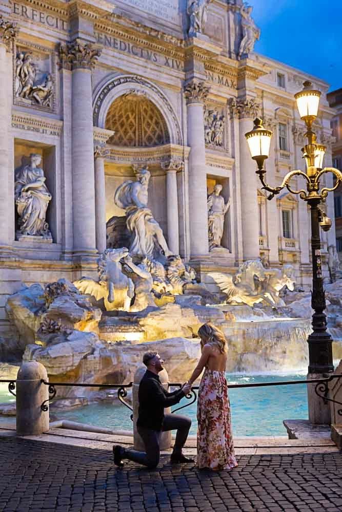 a man kneeling down next to a woman in front of a fountain