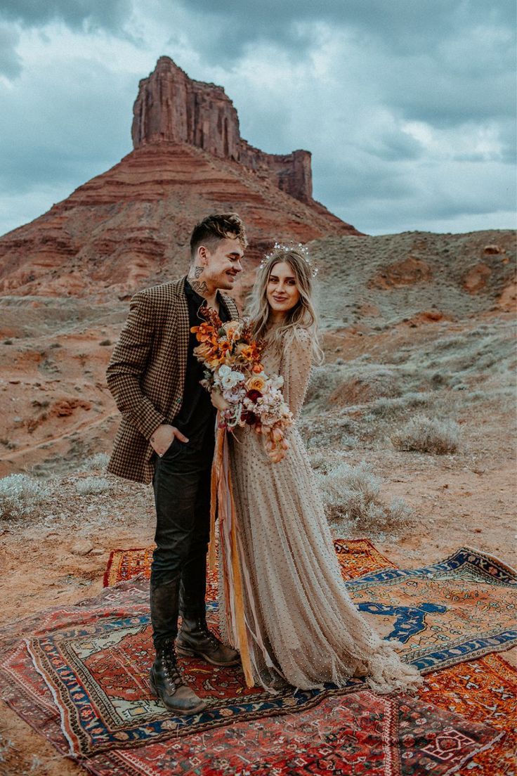 a man and woman standing on top of a rug in the middle of desert with a mountain behind them