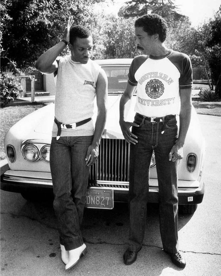 two men standing next to each other in front of a car