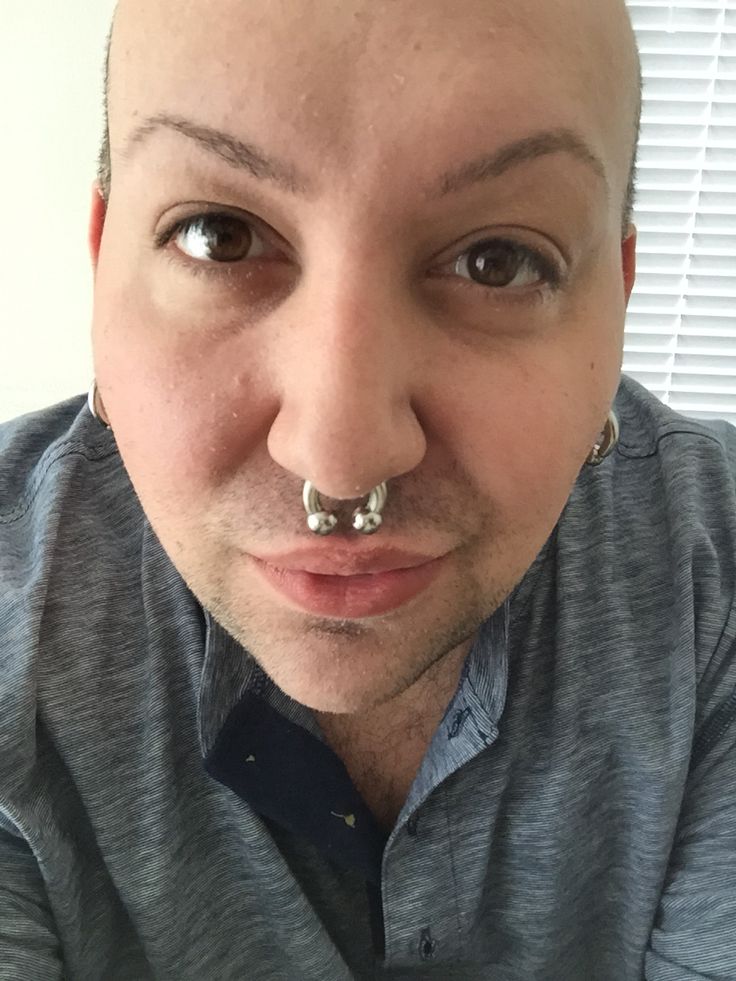 a bald man with piercings on his nose looks at the camera while wearing a gray shirt