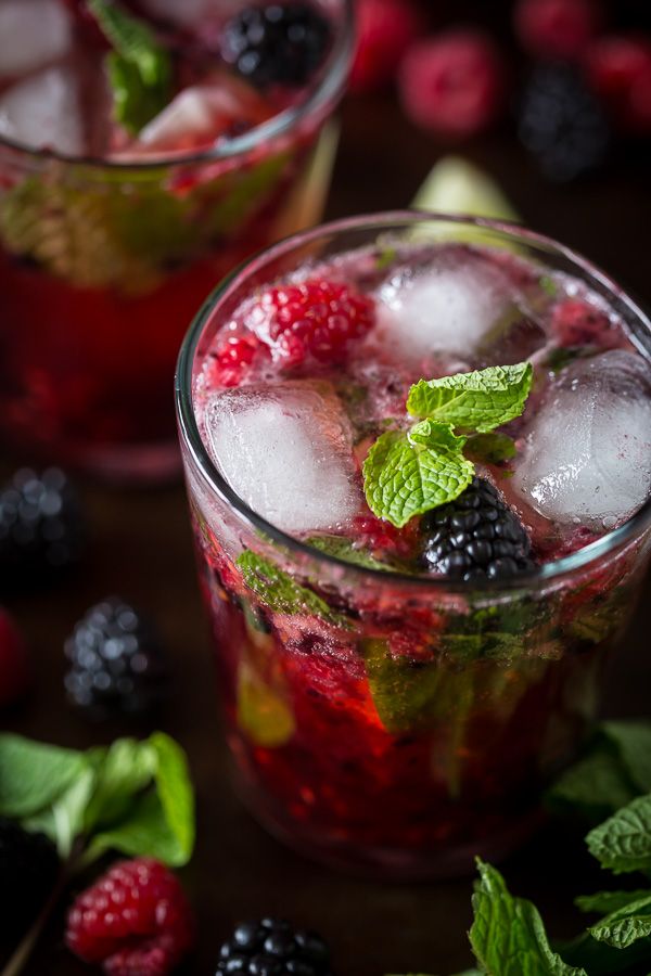 two glasses filled with ice and raspberries on top of a table next to mint leaves