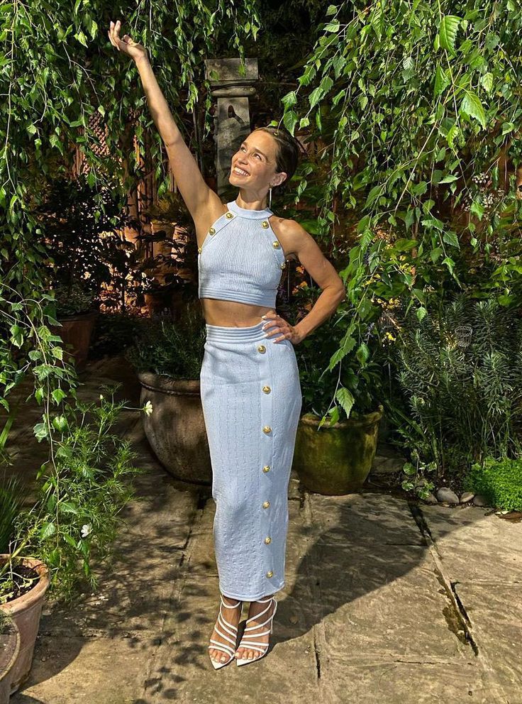 a woman standing in front of some trees and holding her hand up to the sky