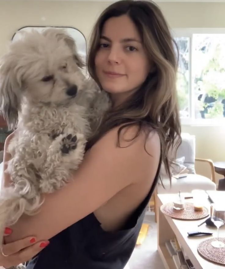 a woman holding a white dog in her arms while standing next to a dining room table