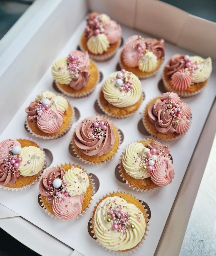 twelve cupcakes with pink and white frosting in a box on a table