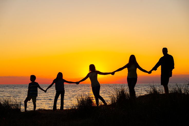 a family holds hands as the sun sets over the water in the background, while silhouetted against an orange and yellow sky