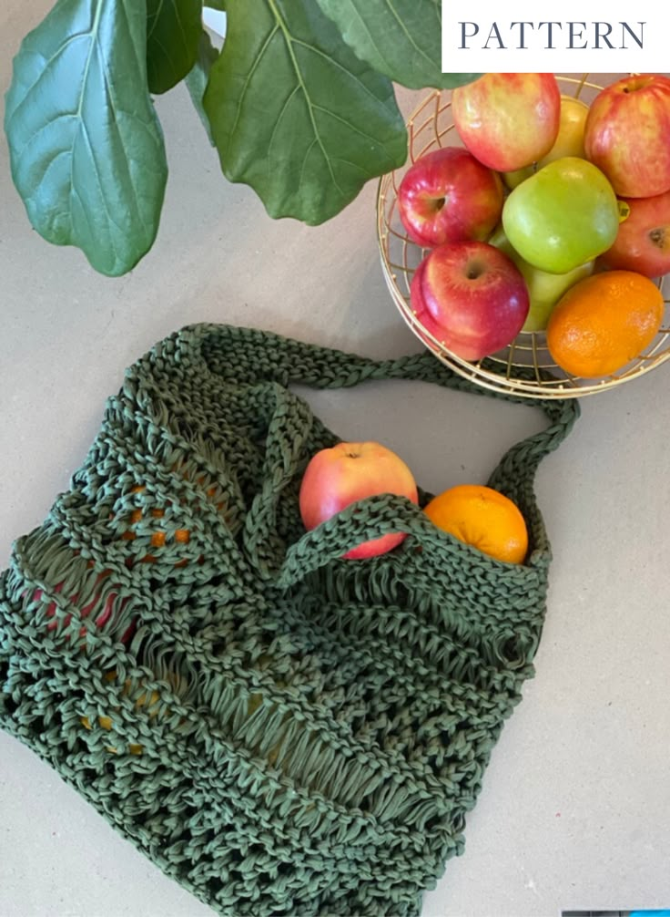 a green crocheted bag with apples and oranges in it next to a basket of fruit