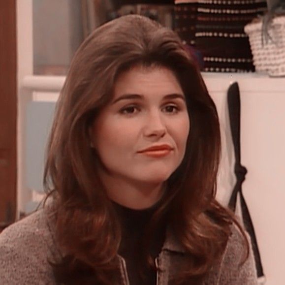 a woman with long brown hair sitting in front of a white refrigerator and looking off to the side