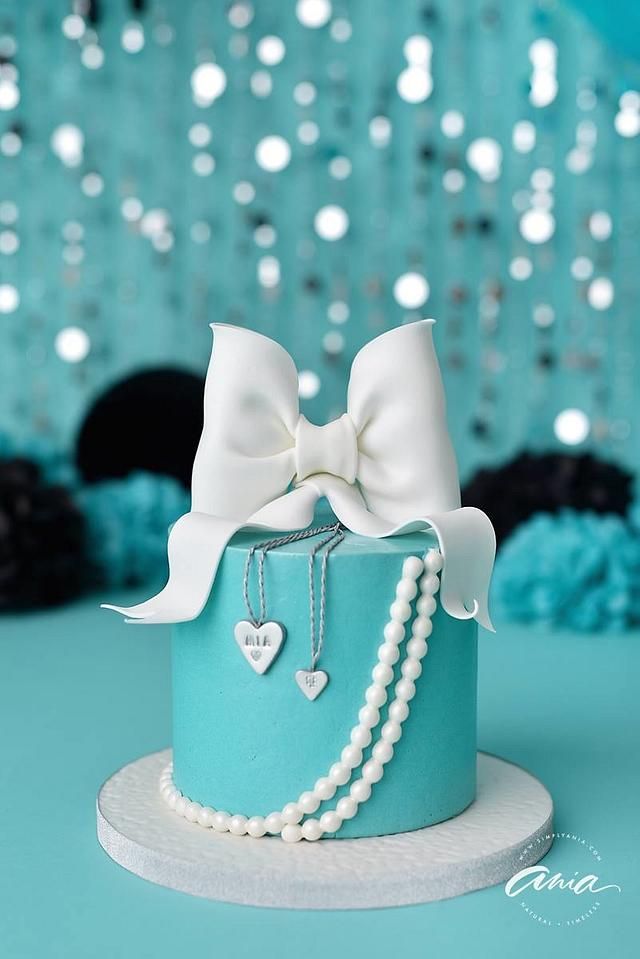 a blue cake with pearls and a white bow on it's top, sitting on a table