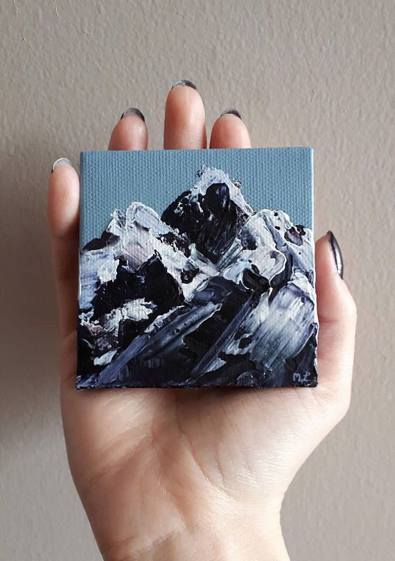 a hand holding up a small painting with mountains in the background