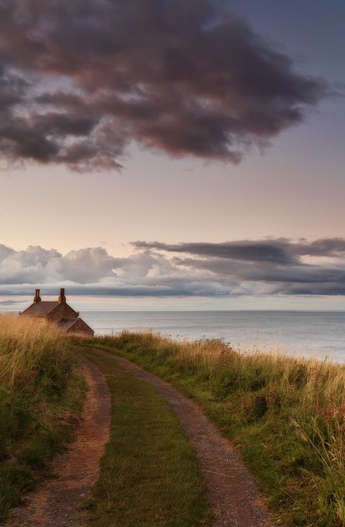 a dirt path leading to a small house on the side of a hill next to the ocean