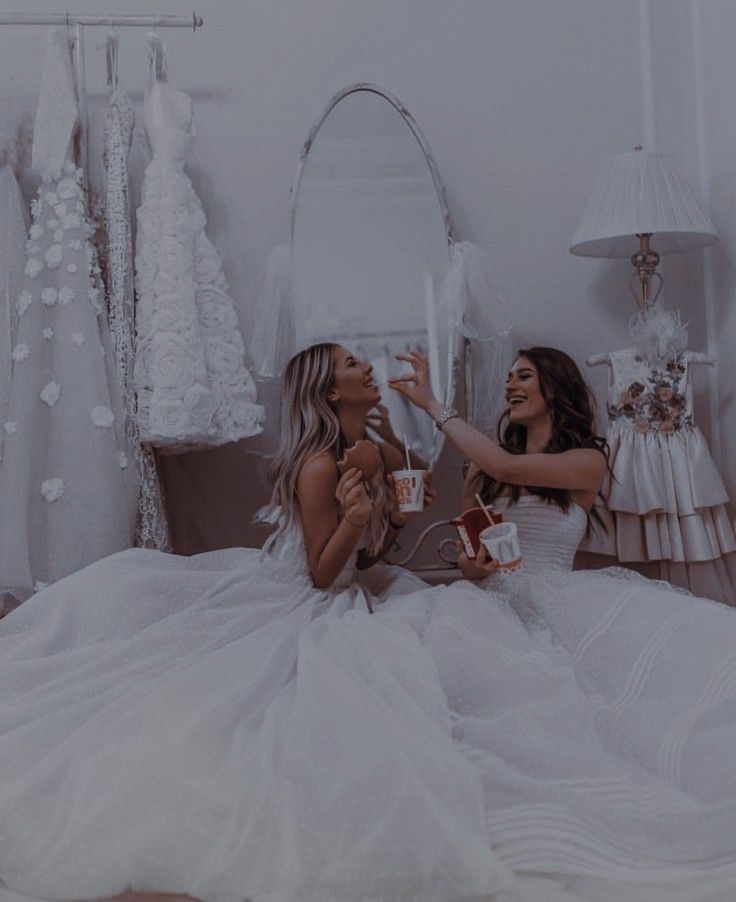 two women in white dresses sitting on a bed and one woman is holding a cup