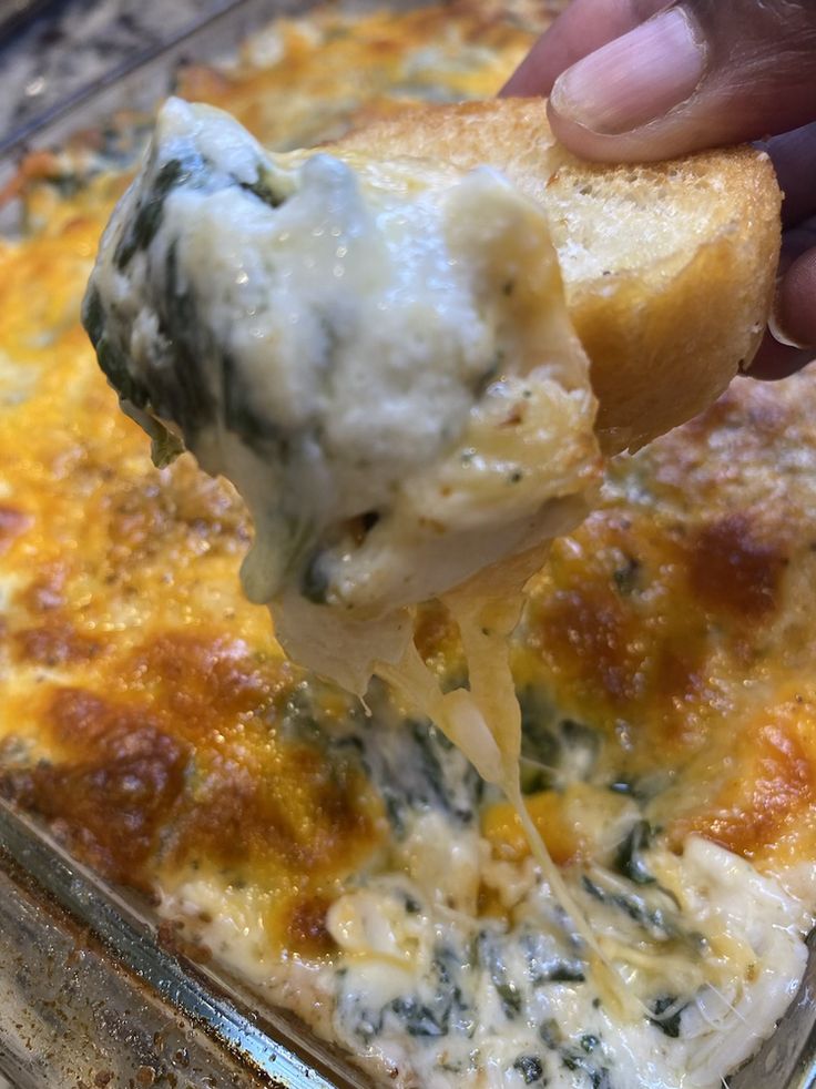 a hand holding a piece of bread with cheese and spinach on it in a casserole dish