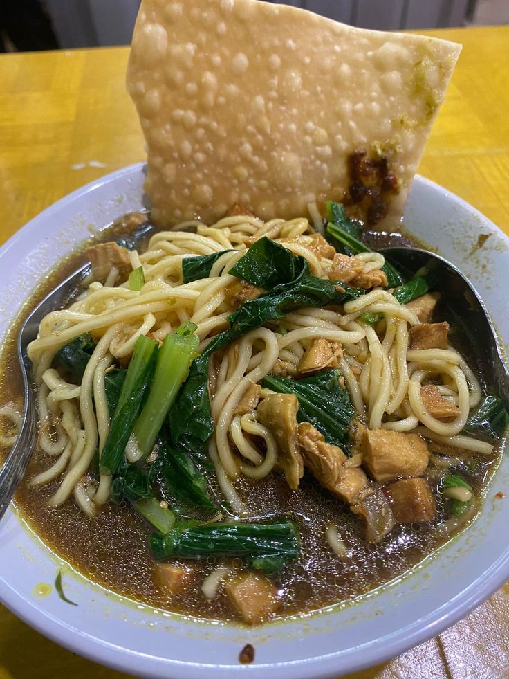 a white bowl filled with noodles and greens next to a tortilla shell on top of a yellow table