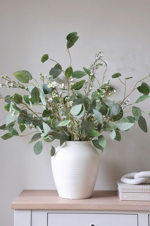 a white vase filled with green leaves on top of a wooden table next to a book