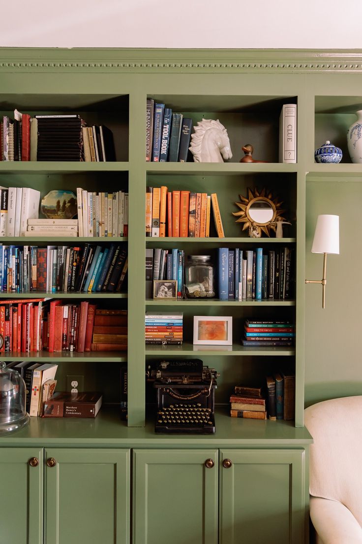 a green bookcase filled with lots of books next to a white chair and lamp