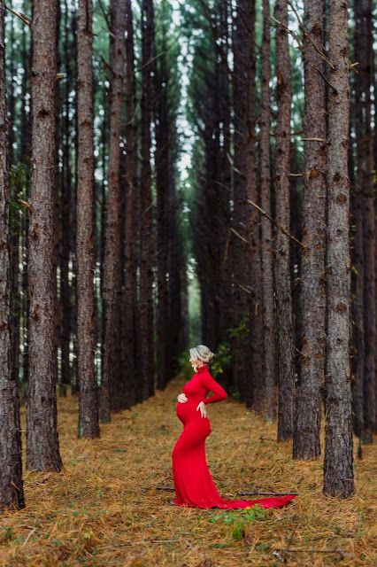 a pregnant woman in a red dress standing in the woods with her hands on her hips