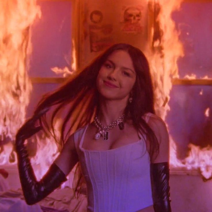 a woman with long hair and leather gloves standing in front of a fire filled wall