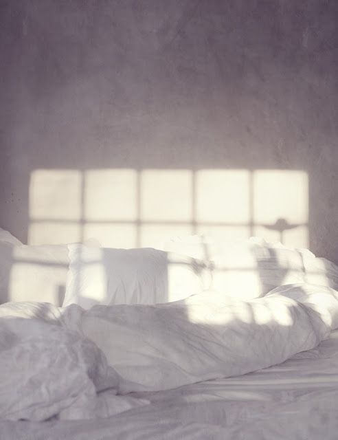 an unmade bed with white sheets and pillows in front of a window that has the sun shining through it