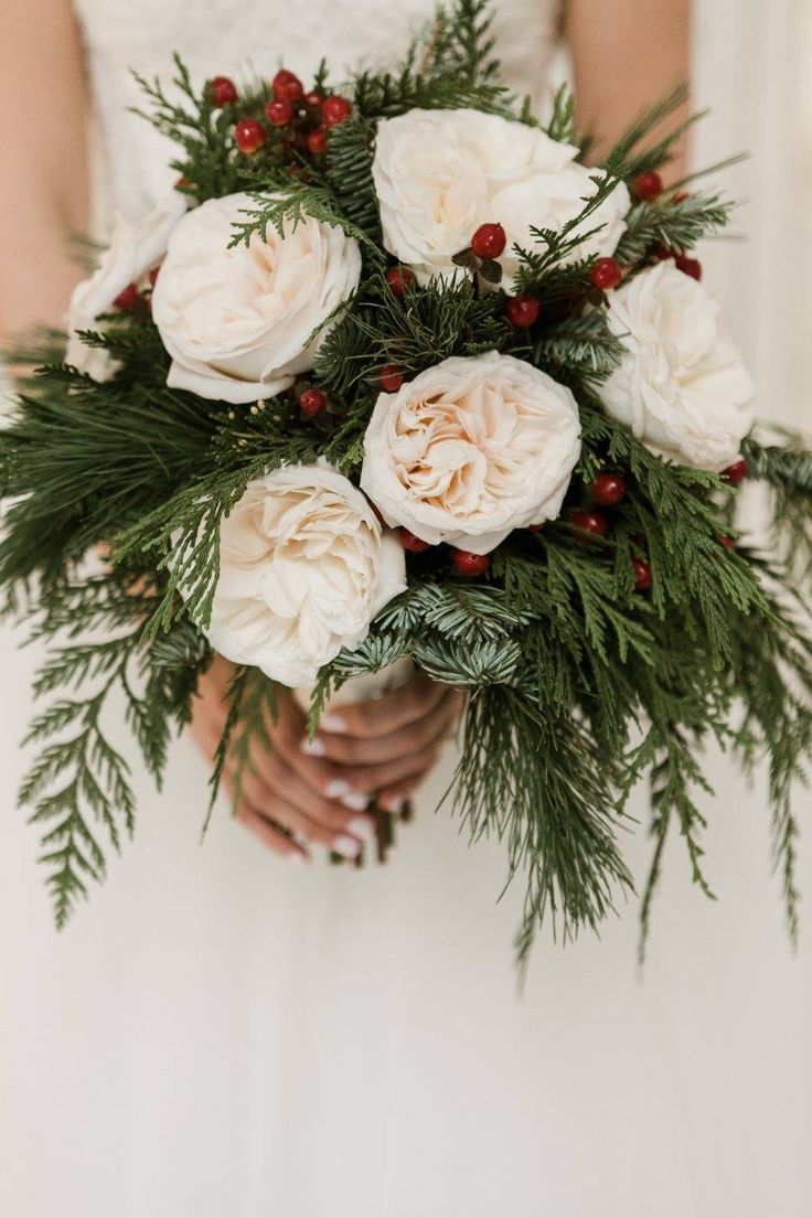 a bride holding a bouquet of white flowers and greenery in her hands with red berries