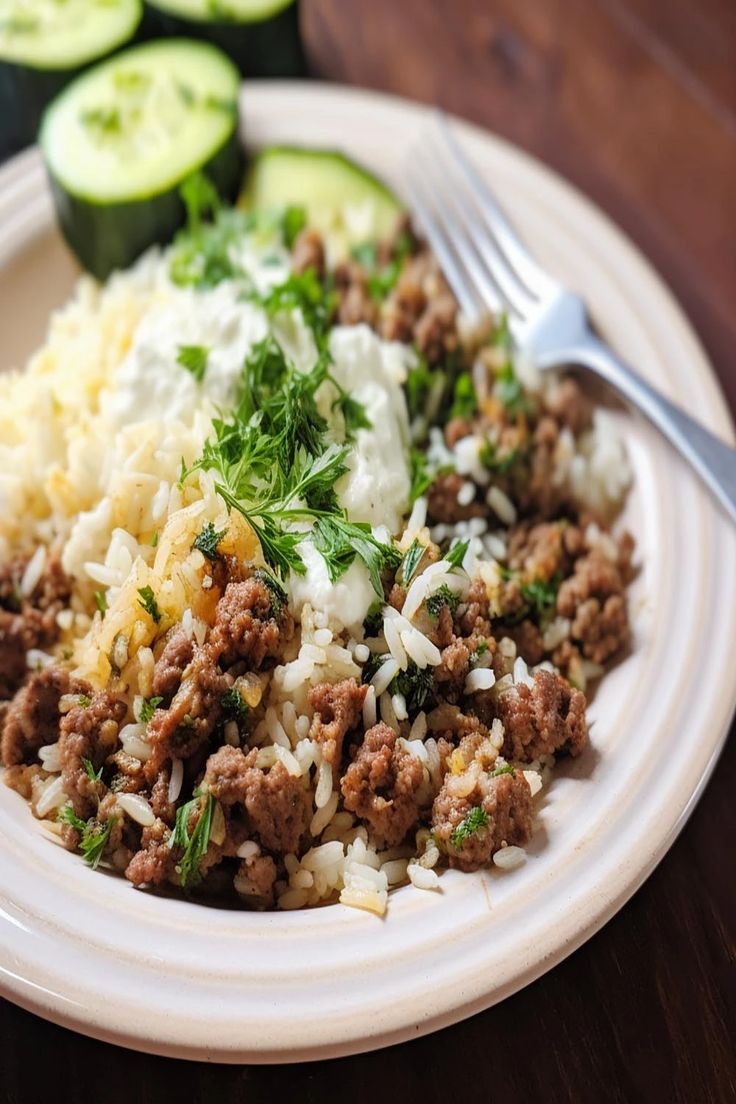 a white plate topped with rice and meat next to cucumber slices on a wooden table