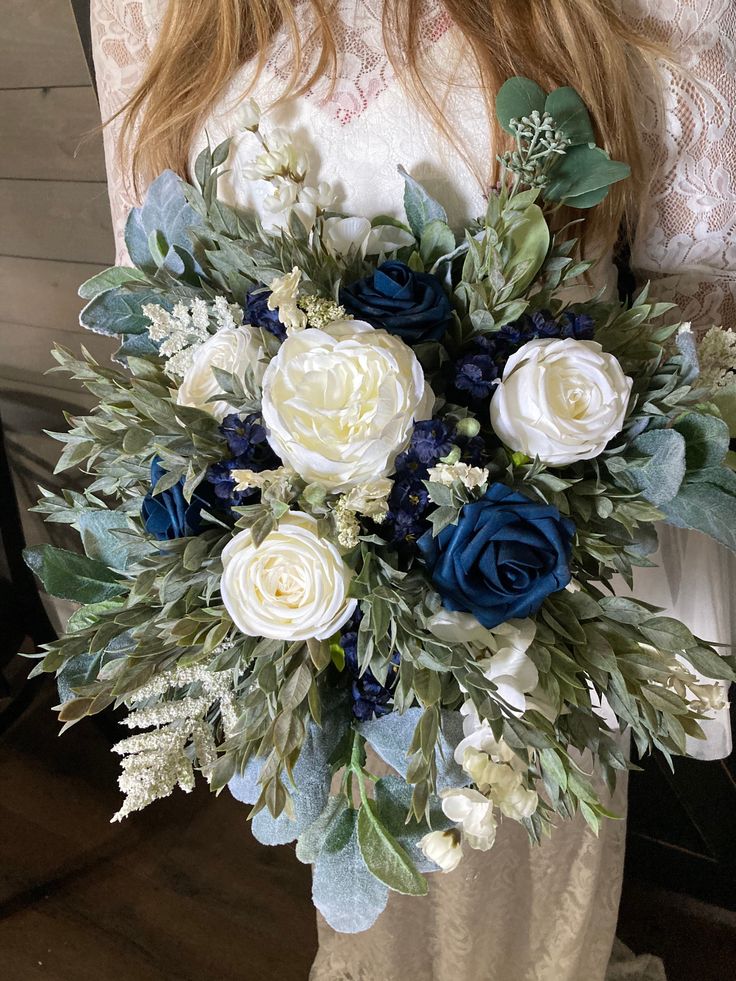 a woman holding a bouquet of white and blue flowers with greenery in her hands