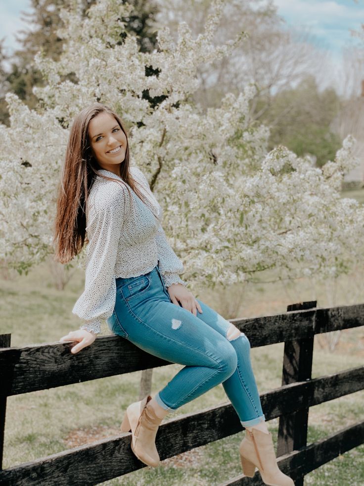 a beautiful young woman sitting on top of a wooden fence next to a flowering tree