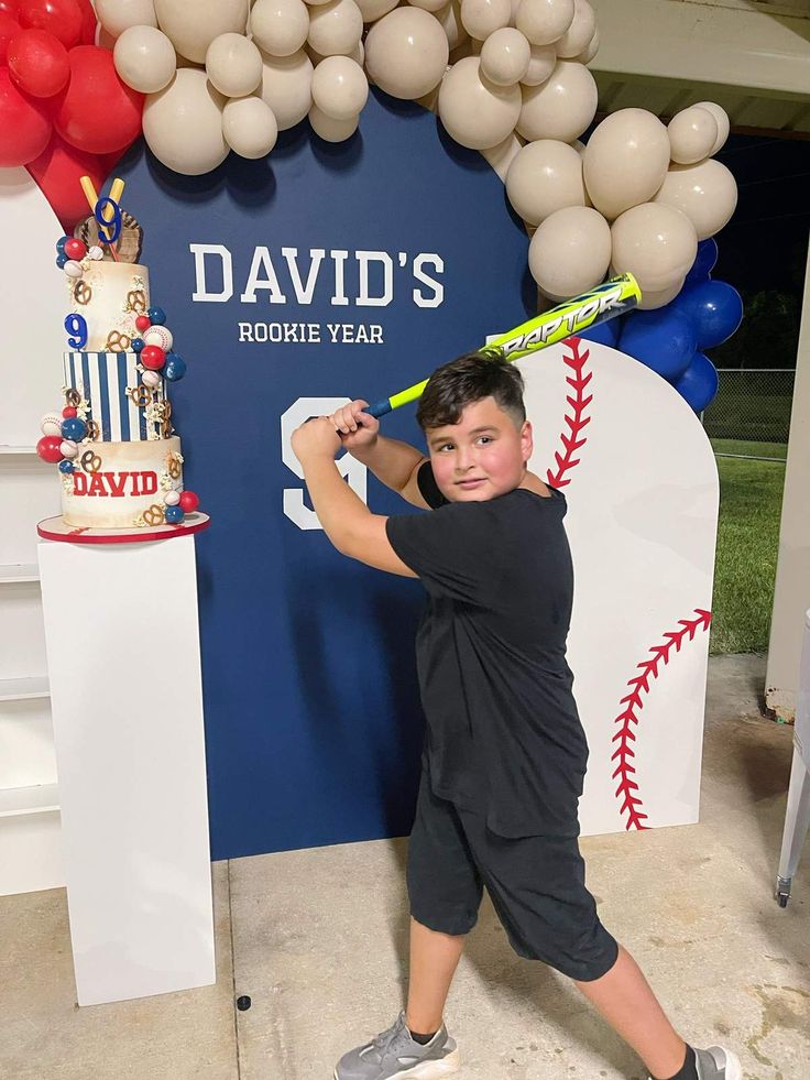 a young boy holding a baseball bat in front of a sign with balloons on it