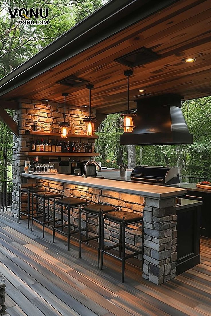 A rustic outdoor bar and kitchen featuring a stone counter, wooden ceiling, and warm lighting. The setup creates a cozy atmosphere, perfect for evening gatherings in a natural, wooded setting. Modern Rustic Outdoor Kitchen, Outside Kitchen With Bar, Bar Area Outdoor, Open Bar Design Outdoor, Modern Outdoor Bar Ideas Backyards, Outdoor Food Setup, Outdoor Entertainment Patio Ideas, Deck With Bar Counter, Pool House Bar Ideas